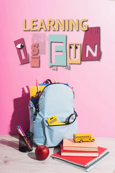 School backpack full of stationery, pen holder, ripe apple, books and toy school bus near learning is fun lettering on pink — Stock Photo