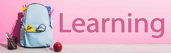 Imagen horizontal de mochila azul con útiles escolares cerca de manzana entera, porta bolígrafos con rotuladores y letras de aprendizaje en rosa - foto de stock