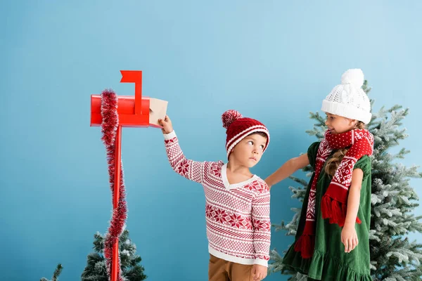 Boy in hat and sweater putting envelope in mailbox near sister in winter outfit isolated on blue — Stock Photo