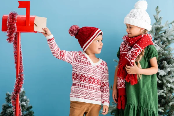 Boy in knitted sweater putting envelope in mailbox and looking at sister in winter outfit isolated on blue — Stock Photo