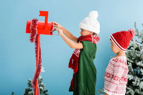 Criança em roupa de inverno colocando envelope na caixa de correio perto do irmão isolado em azul — Fotografia de Stock