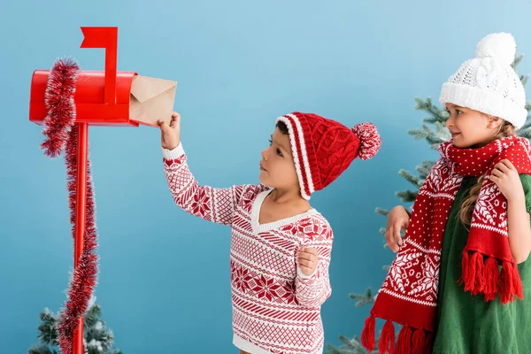 Boy in knitted sweater and hat putting envelope in mailbox near sister in winter outfit isolated on blue — Stock Photo