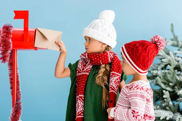 Дівчина в зимовому вбранні кладе конверт в поштову скриньку біля брата ізольовано на синьому — стокове фото