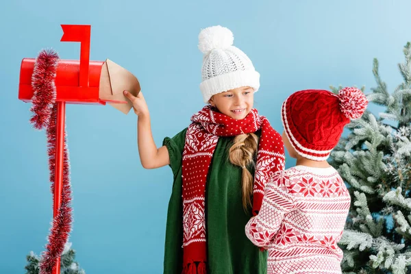 Girl in winter outfit putting envelope in mailbox and looking at brother isolated on blue — Stock Photo