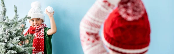 Foyer sélectif de l'enfant dans le chapeau et l'écharpe tenant boule de neige tout en jouant avec son frère sur bleu, plan panoramique — Photo de stock