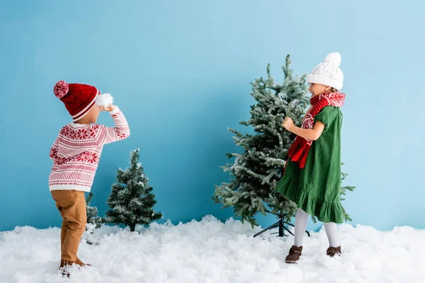 Kinder im Winteroutfit spielen Schneebälle in der Nähe von Weihnachtsbäumen auf blau — Stockfoto