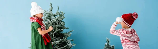 Plano panorámico de los niños en traje de invierno jugando bolas de nieve cerca de árboles de Navidad en azul - foto de stock