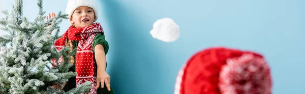 Récolte panoramique de fille en écharpe jetant boule de neige près de frère en chapeau sur bleu — Photo de stock
