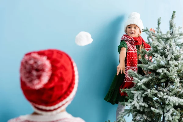 Selective focus of girl in hat and scarf throwing snowball near brother on blue — Stock Photo