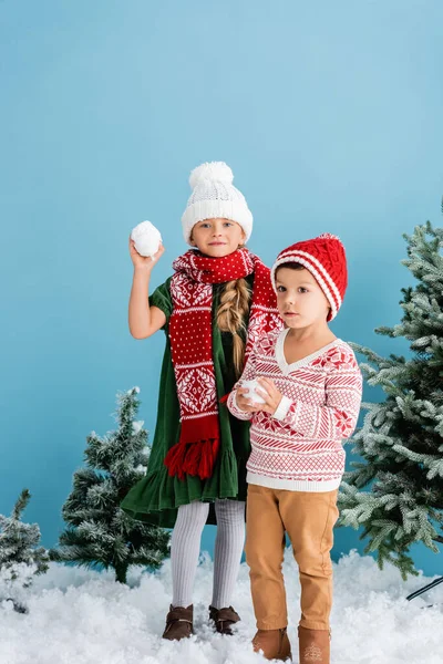 Kids in winter outfit holding snowballs near christmas trees on blue — Stock Photo