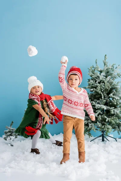 Kinder im Winteroutfit spielen Schneebälle in der Nähe von Weihnachtsbäumen auf blau — Stockfoto