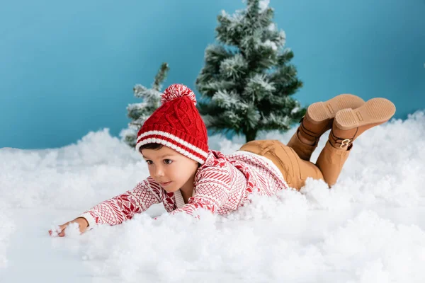Boy in winter outfit lying on white snow near christmas trees on blue — Stock Photo