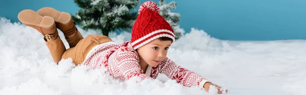 Panoramic concept of boy in winter outfit lying on white snow near christmas trees on blue — Stock Photo