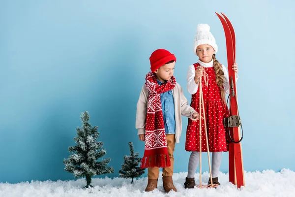 Kind mit Mütze und Schal zeigt mit dem Finger auf Schwester, die Skistöcke und Skier hält, während sie auf blau steht — Stockfoto