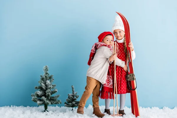Хлопчик в капелюсі і шарфі обіймає сестру з лижними полюсами і лижами, стоячи на синьому — стокове фото