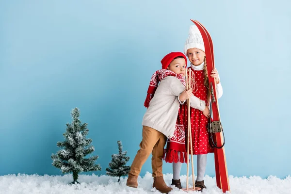 Junge mit Mütze und Schal umarmt Schwester mit Skistöcken und Skiern im Stehen auf blau — Stockfoto