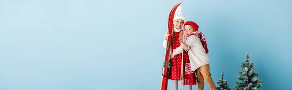 Concepto panorámico de niño en traje de invierno abrazando hermana con bastones de esquí y esquís en azul - foto de stock
