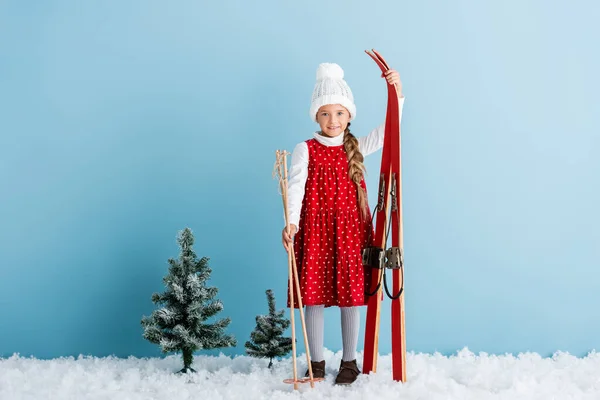 Ребенок в зимнем наряде стоит на снегу с лыжными палками и лыжами на синем — стоковое фото