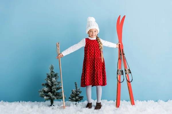 Kind im Winteroutfit auf Schnee stehend mit Skistöcken und Skiern auf blau — Stockfoto