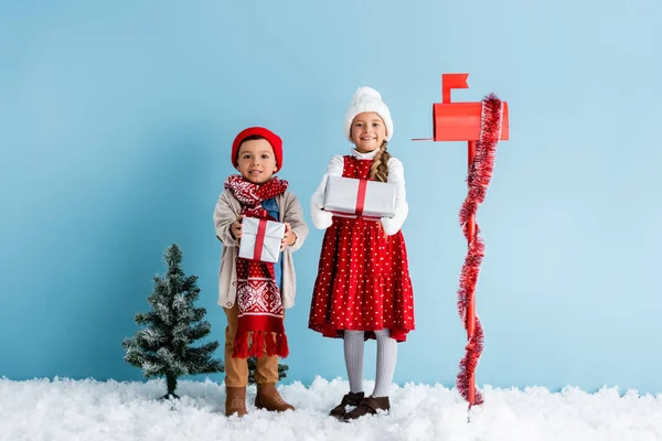 Crianças em roupa de inverno em pé na neve e segurando presentes perto de caixa de correio e abeto em azul — Fotografia de Stock