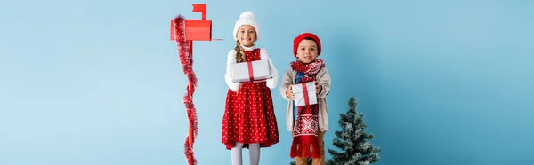 Plan panoramique des enfants en tenue d'hiver tenant des cadeaux près de la boîte aux lettres et sapin sur bleu — Photo de stock