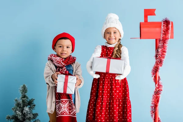 Enfants en chapeaux et tenue d'hiver tenant des cadeaux près de la boîte aux lettres et sapin sur bleu — Photo de stock