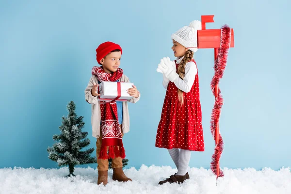 Junge im Winteroutfit hält Geschenk neben Schwester und Briefkasten auf blauem Grund — Stockfoto