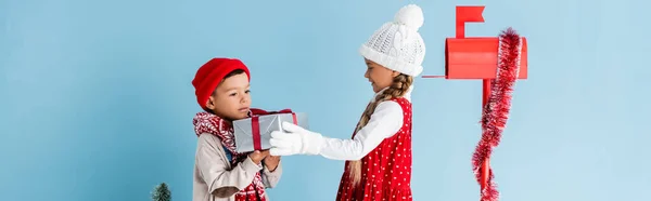 Panoramisches Konzept des Jungen im winterlichen Outfit mit Geschenk in der Nähe von Schwester und Briefkasten isoliert auf blau — Stockfoto