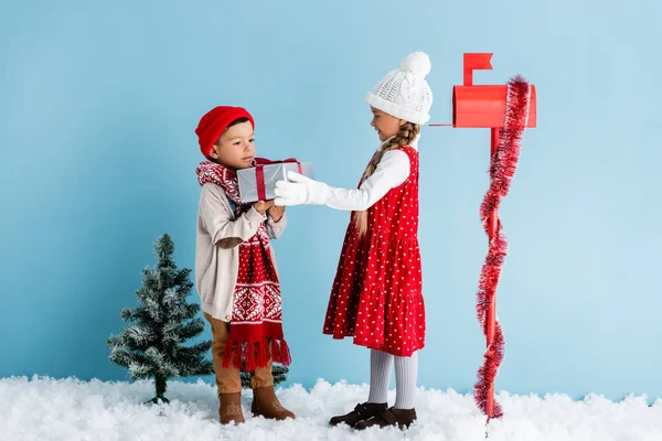 Garçon en tenue d'hiver tenant présent près de soeur dans chapeau et boîte aux lettres sur bleu — Photo de stock