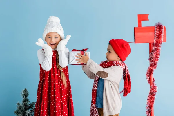 Junge im Winteroutfit hält Geschenk neben aufgeregter Schwester und Briefkasten isoliert auf blauem Grund — Stockfoto