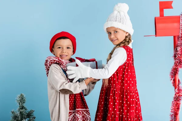 Sister and brother in winter outfit holding present near mailbox on blue — Stock Photo