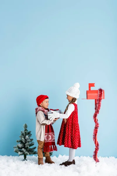 Soeur et frère en tenue d'hiver se regardant tout en tenant présent près de la boîte aux lettres sur bleu — Photo de stock