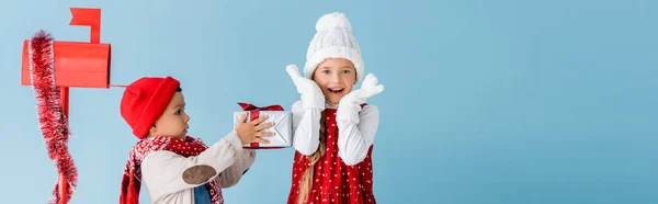 Horizontale Ernte von Junge im Winteroutfit hält Geschenk in der Nähe aufgeregter Schwester und Briefkasten isoliert auf blau — Stockfoto