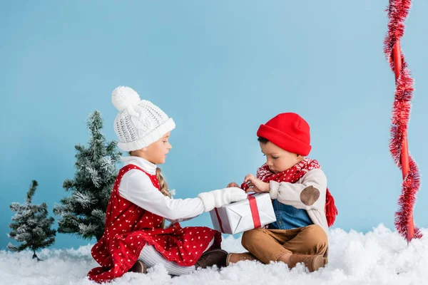 Boy and girl in winter outfit sitting on snow and touching present isolated on blue — Stock Photo