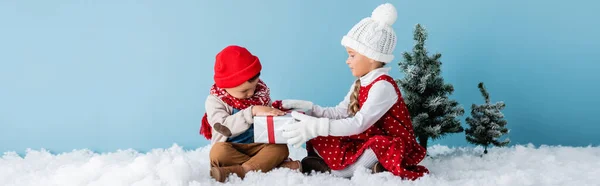 Panoramic shot of sister and brother in winter outfit sitting on snow and touching present on blue — Stock Photo