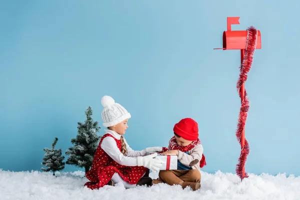 Kinder im Winteroutfit sitzen auf Schnee und rühren Geschenk in der Nähe Briefkasten auf blau — Stockfoto