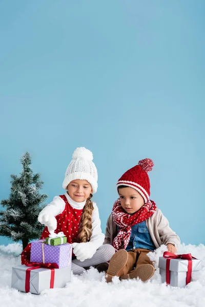 Niños en traje de invierno sentados en la nieve y mirando regalos cerca de abeto aislado en azul - foto de stock