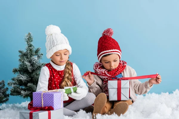 Kinder im Winteroutfit sitzen auf Schnee in der Nähe von Tannen und halten Geschenke isoliert auf blauem Grund. — Stockfoto