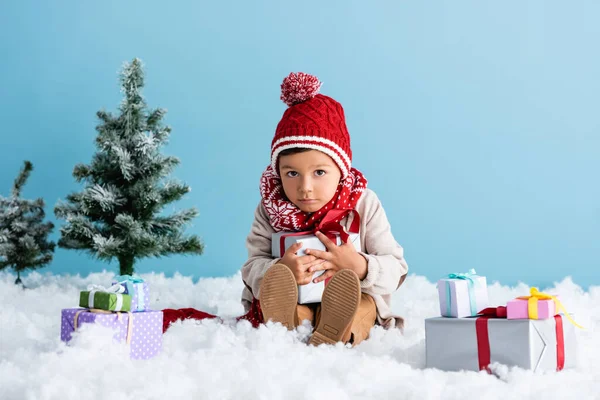 Boy in hat and winter outfit sitting on snow and holding present near christmas trees isolated on blue — Stock Photo