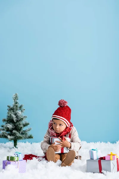 Menino de chapéu e roupa de inverno sentado na neve e segurando caixa de presente perto de árvores de natal isolado em azul — Fotografia de Stock