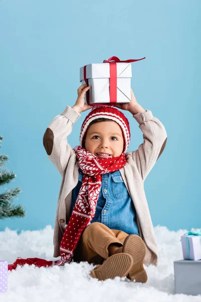 Bambino in cappello e vestito invernale seduto sulla neve e tenendo presente sopra la testa isolato su blu — Foto stock