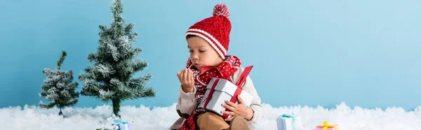 Conceito panorâmico de menino de chapéu e roupa de inverno sentado na neve e segurando presente enquanto soprando na mão isolado em azul — Fotografia de Stock