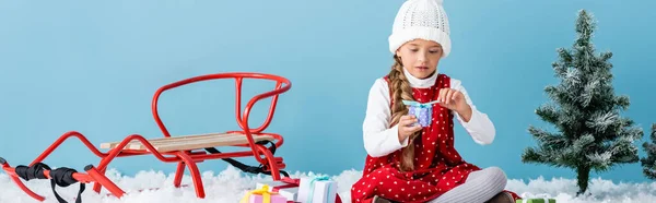 Panoramic shot of kid in hat and winter outfit sitting on snow and holding present near sleigh isolated on blue — Stock Photo