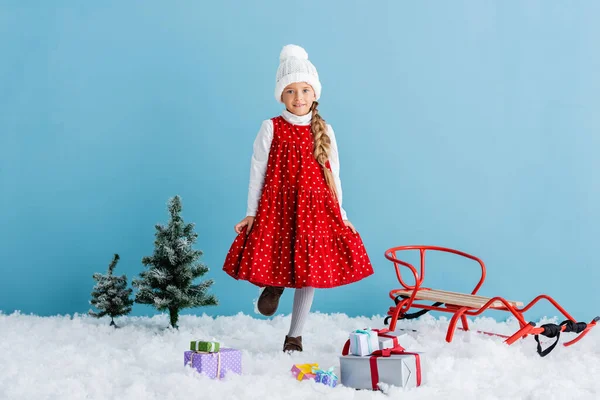 Kid in hat and winter outfit standing on snow near presents and sleigh isolated on blue — Stock Photo