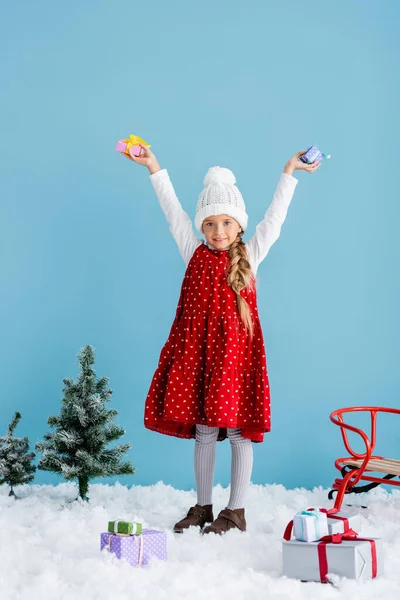 Kid in hat and winter outfit standing on snow and holding presents above head near sleigh isolated on blue — Stock Photo