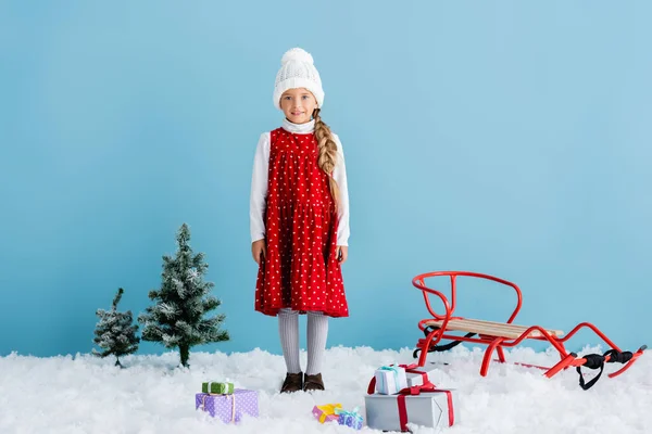 Girl in hat and winter outfit standing on snow near presents and sleigh isolated on blue — Stock Photo