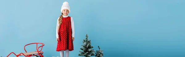 Cultivo panorámico de chica en sombrero y traje de invierno de pie en cerca del trineo y pinos aislados en azul - foto de stock