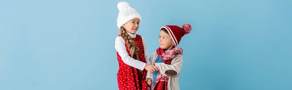 Cultivo panorámico de niño en sombrero cogido de la mano con la hermana aislada en azul - foto de stock