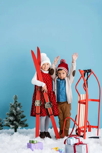 Girl in winter outfit holding skis near brother with hands above head standing near sleight and presents on snow isolated on blue — Stock Photo