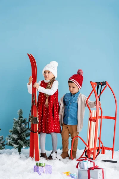 Girl in winter outfit holding skis near boy standing near sleight and presents on snow isolated on blue — Stock Photo